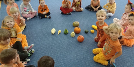 dzieci siedzą na dywanie w pomarańczowych ubraniach. Na środku dywanu poukładane są dynie