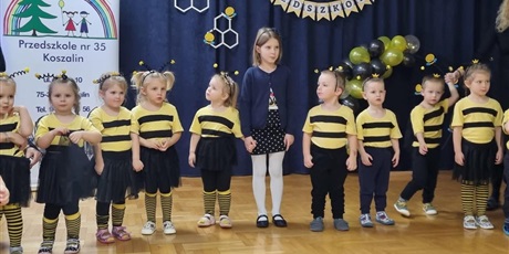 dzieci ubrane w żółto- czarne stroje stoją w szeregu. Za nimi dekoracja na granatowym tle- napis " pasowanie na przedszkolaka"