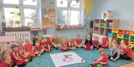 Grupa dzieci, chłopcy i dziewczynki, ubrani na czerwono, siedzący w półkolu na dywanie. W środku przed nimi leży plansza do kodowania z zakodowanym jabłkiem.
