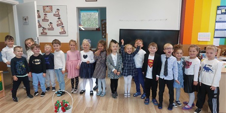 Grupa dzieci, chłopcy i dziewczynki, ubrani elegancko, w sukienki, koszule, stoją w rzędzie, uśmiechając się. Przed nimi stoi biały koszyk z czerwonymi kwiatkami z bibuły.