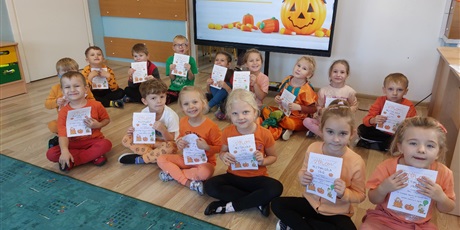 Grupa dzieci, chłopcy i dziewczynki, siedzą na podłodze przed ekranem z wyświetlonym napisem Dzień dyni. Dzieci są ubrane na pomarańczowo i trzymają w rękach dyplomy.