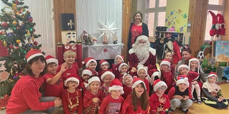 Grupa dzieci, chłopcy i dziewczynki, wszyscy ubrani na czerwono i w czapkach mikołajowych, siedzą przed dekoracją kominka. Z nimi siedzi Mikołaj, trzy kobiety, szatynki, także ubrane na czerwono.