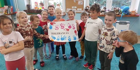 Grupa dzieci, chłopcy i dziewczynki stoją na turkusowym dywanie i trzymają plakat z odciśniętymi czerwoną i niebieską farbą plakat. Na środku plakatu jest napis tolerancja.