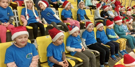 Grupa dzieci, chłopcy i dziewczynki, siedzą na trybunach. Dzieci są ubrane w niebieskie koszulki z Logo Przedszkola nr 35 i czerwone czapki mikołajkowe.