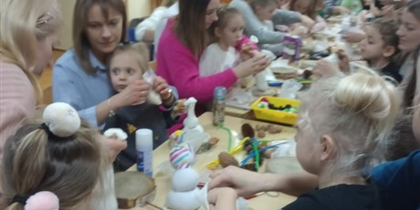 Grupa dzieci i ich rodzice, siedzą przy długim stole, połączonym z wielu mniejszych i tworzą dekoracje. Dzieci siedzą na kolanach u rodziców i razem z rodzicami są pochłonięte zadaniem plastycznym.