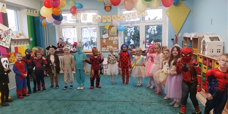 Grupa dzieci, chłopcy i dziewczynki, stoją na turkusowym dywanie w półkolu. Dzieci są przebrane za różne postacie: superbohater, pirat, królik, księżniczka, jednorożec.