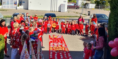 Grupa dzieci, chłopcy i dziewczynki, wśród nich kobiety, wszyscy ubrani na czerwono, stoją w kole, na środku leży czerwony dywan ozdobiony biedronkami, pszczółkami, kotkami, motylami, misiami i sowami