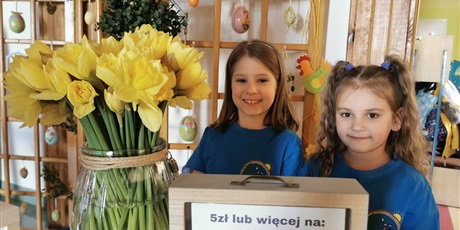 na pierwszym planie skarbonka na zbiórkę pieniędzy, obok wazon z żółtymi kwiatami- żonkilami, Na drugim planie dwie dziewczynki w niebieskich koszulkach.