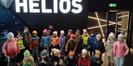 Grupa dzieci, chłopcy i dziewczynki, szesnaścioro stoi, sześcioro kuca pod napisem Helios. Dzieci, ubrane w kurtki i czapki,  uśmiechają się do zdjęcia.
