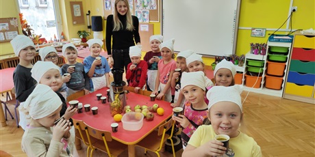 Grupa dzieci, chłopcy i dziewczynki, z białymi czepkami na głowach, stoją wokół stołu, na którym stoi sokowirówka, wiele owoców i kubeczki z sokiem. Za nimi stoi kobieta, blondynka.