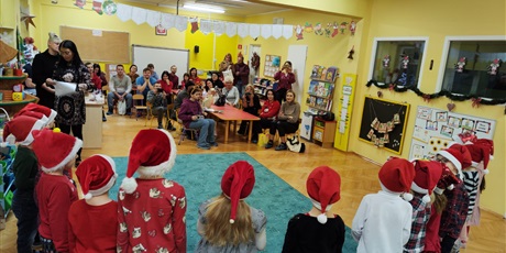 Grupa dzieci, chłopcy i dziewczynki, stoją w półkolu na turkusowym dywanie i śpiewają piosenkę rodzicom, stojącym lub siedzącym przy czerwonych stolikach. Dzieci mają na sobie czerwone czapki.