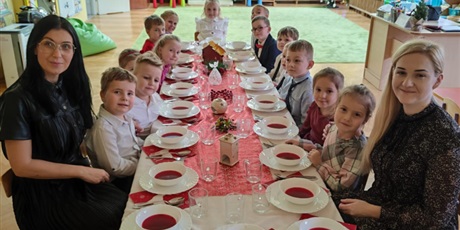 Grupa dzieci, chłopcy i dziewczynki, elegancko ubrani, dziewczynki w sukienki, chłopcy w koszule, siedzą przy stole, odświętnie ozdobionym. Przed każdym z dzieci stoi zupa, barszcz czerwony.