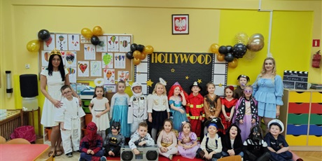 Grupa dzieci, chłopcy i dziewczynki, przebrani w różne stroje: syrenka, policjant, pirat, superbohaterowie, księżniczki, aniołki stoją lub kucają, przed ścianą z napisem Hollywood, obok stoją kobiety.