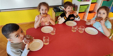 Czworo dzieci, dwóch chłopców, brunetów, dwie dziewczynki, szatynki, siedzą przy stoliku, przed każdym dzieckiem leży talerz i stoi szklanka z wodą. Przy twarzach dzieci trzymają pączki z lukrem.