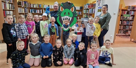 Grupa dzieci, chłopcy i dziewczynki, stoją lub kucają w bibliotece. Za nimi stoją dwie kobiety, jedna brunetka a druga blondynka, trzymająca nad jednym z dzieci wielką głowę zielonego dinozaura.