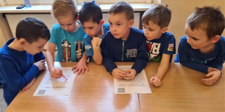 Sześciu chłopców, ubranych na niebiesko, stoi i opiera się o złączone ławki szkolne, na których są dwie zagadki matematyczne. Jeden z chłopców, blondyn wykonuje zadanie.