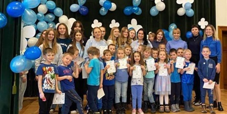 Grupa dzieci, chłopcy i dziewczynki, ubrani na niebiesko stoją i uśmiechają się do zdjęcia, z tyłu stoją cztery kobiety, dwie blondynki, dwie brunetki.