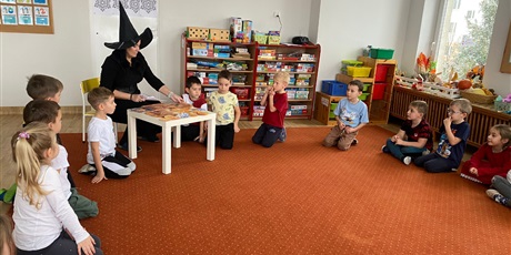 Grupa dzieci, chłopcy i dziewczynki, siedzą w kole na pomarańczowym dywanie. Między nimi kobieta, brunetka, ubrana na czarno, w czarnym kapeluszu czarownicy, przed nią stół, na którym leżą kartki.