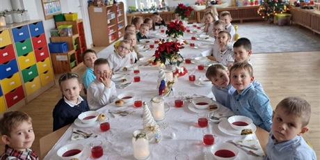 Grupa dzieci, chłopcy ubrani w koszule i dziewczynki ubrane w sukienki lub spódniczki, siedzą przy długim stole, nakrytym białym obrusem. Przed każdym z dzieci stoi zupa - barszcz czerwony.