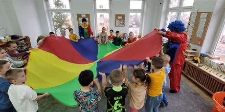 Grupa dzieci, chłopcy i dziewczynki a także dwie kobiety w stroju klaunów, stoją w kole na szarym dywanie, trzymając kolorową chustę Klanzy.