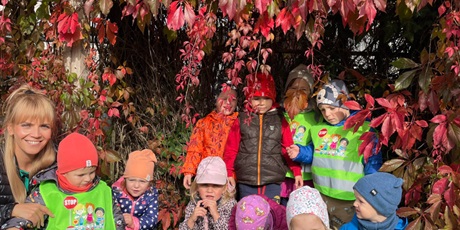 dzieci ustawione w dwóch rzędach w towarzystwie pani, chowające się pod czerwonymi liśćmi pnącego się, czerwonego krzewu