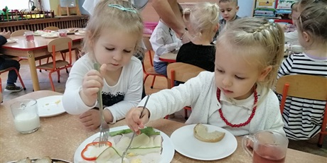 dziewczynki ubrane na biało, samodzielnie przygotowujące sobie kanapkę. Na talerzyku przed nimi: chleb, wędlina, ser, jajka, papryka, sałata