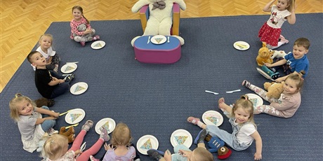 dzieci siedzące w półkolu, przed nimi miś siedzący przy stoliku. Przed dziecmi i przed dziećmi leżą talerzyki oraz sztućce