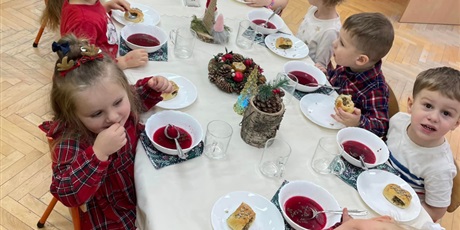 dzieci w odświętnych strojach siedzą przy świąteczym stole nakrytym białym obrusem. Na stole świąteczne dekoracje i talerzyki z czerwonym barszczem
