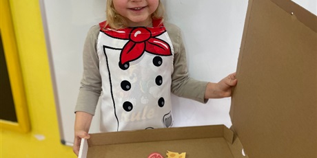 dziewczynka ubrana w strój kucharza, trzymająca karton z pizzą wykonaną z papierowych elementów