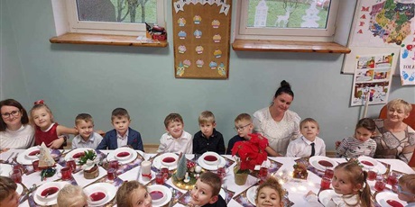 Grupa dzieci, chłopcy i dziewczynki, ubrani elegancko, dziewczynki w sukienki, chłopcy w koszule, siedzą przy świątecznie ozdobionym stole. Przed każdym z dzieci stoi talerz z barszczem czerwonym.